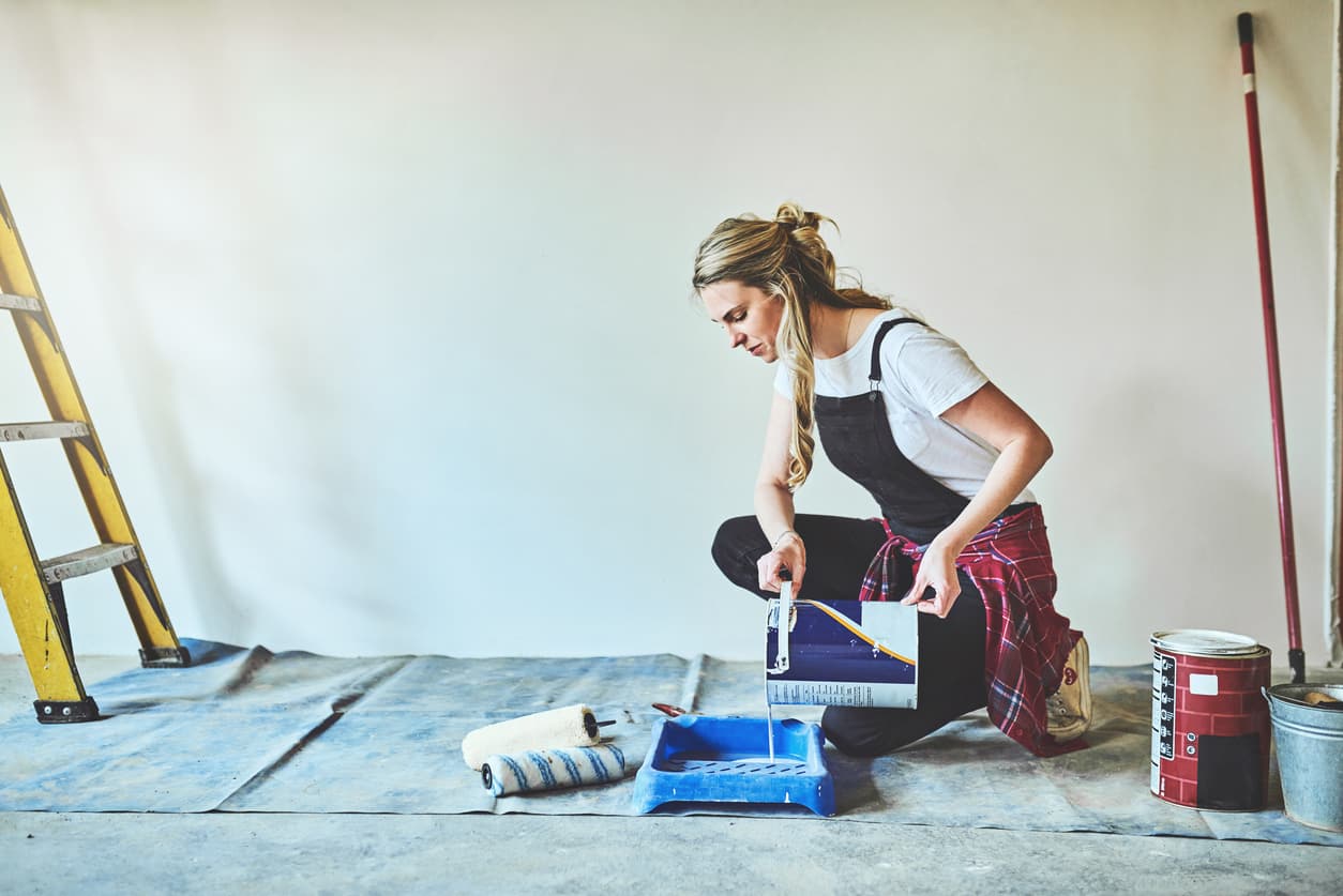 A decorator kneeling on a dust sheet, pouring paint into a roller tray, with a stepladder and extension pole in a room prepped for painting.