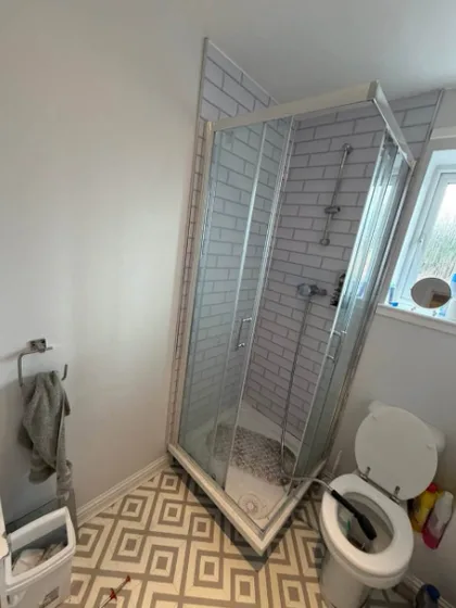 Small bathroom before refit: corner shower cubicle with chrome sliding doors and white metro tiles, next to a toilet, on a diamond-pattern vinyl floor.